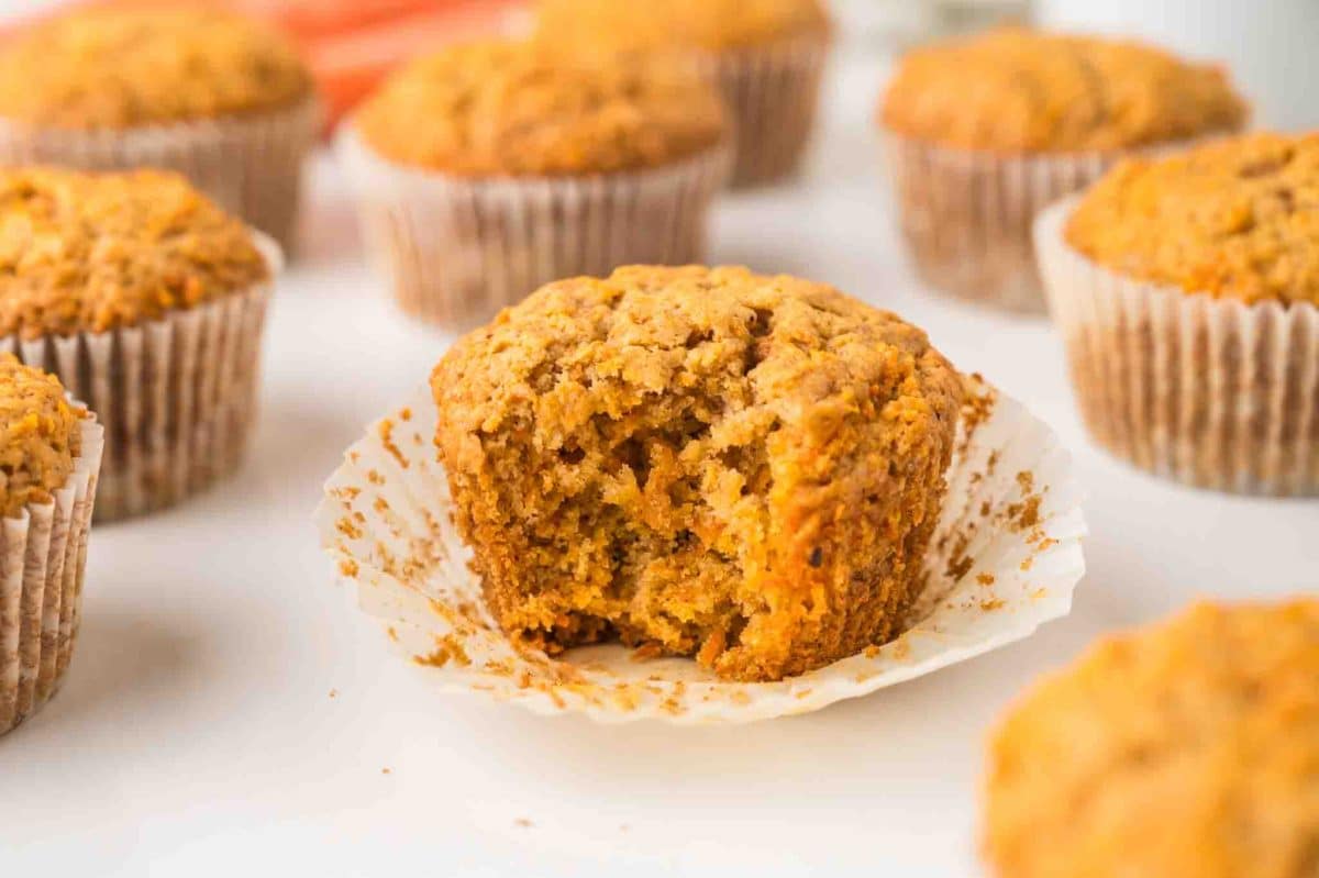 Sideview of carrot cake breakfast muffins sitting on a counter. The middle one is sitting on a muffin paper with a bite taken out of it showing a moist texture with visible spices and carrot pieces. Hostess At Heart