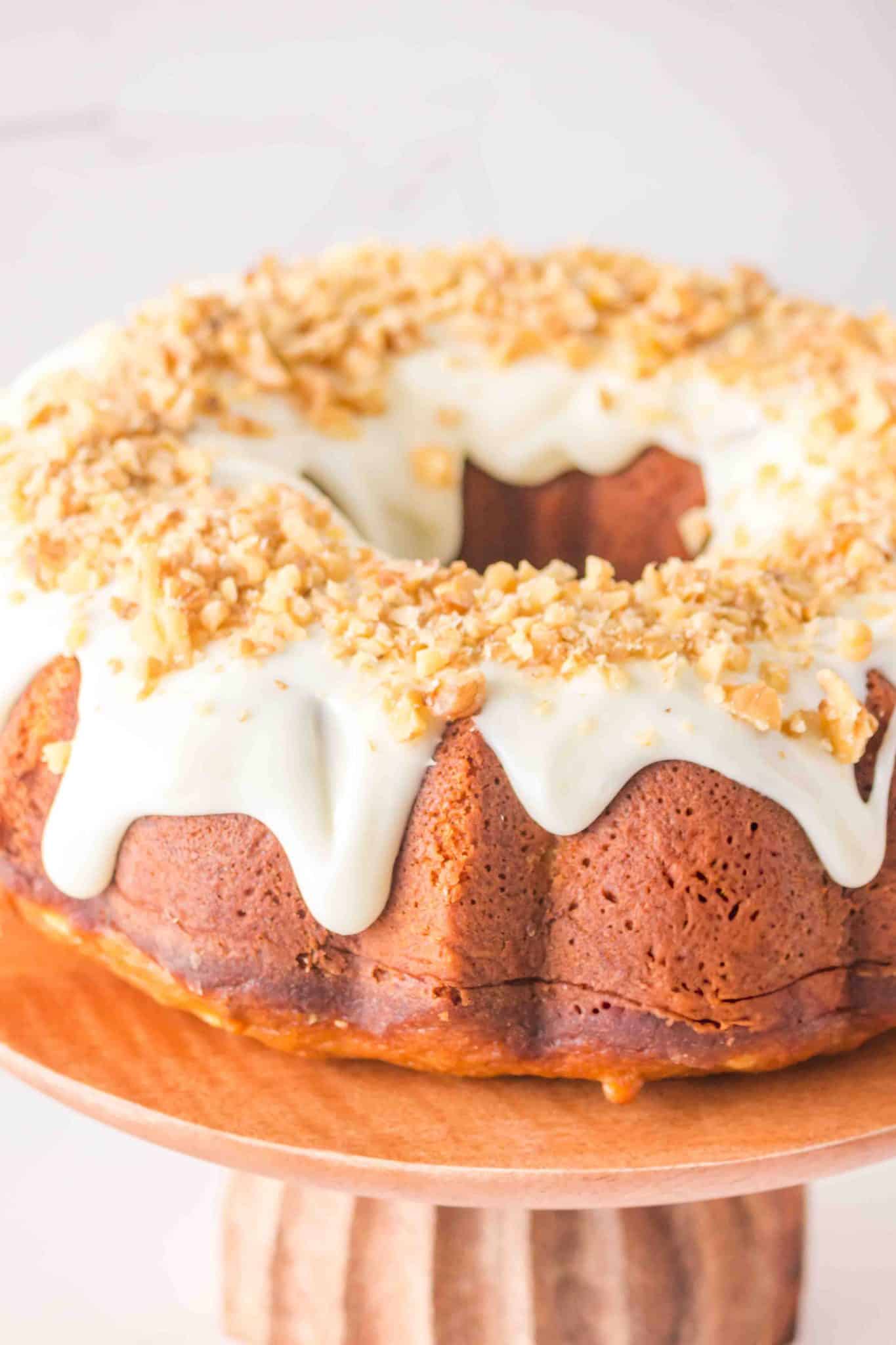 Sideview of a golden brown carrot bundt cake glazed with cream cheese frosting and crushed walnuts on a wooden cake stand. Hostess At Heart