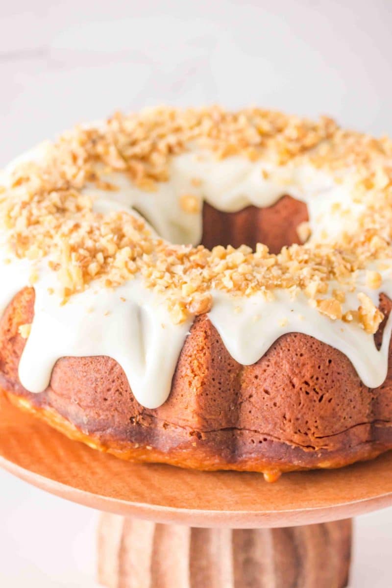 Sideview of a golden brown carrot bundt cake glazed with cream cheese frosting and crushed walnuts on a wooden cake stand. Hostess At Heart