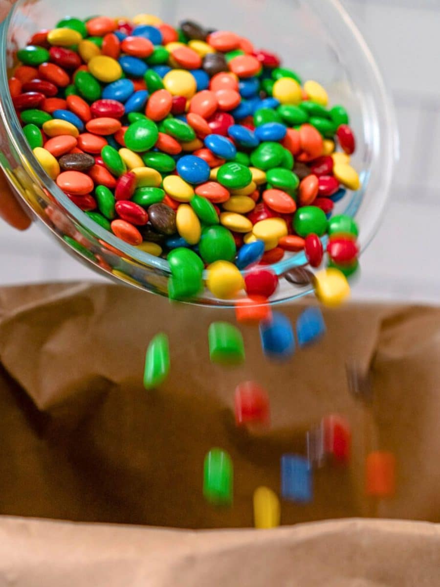 Colorful M&M's being poured into a brown paper bag of peanuts and Crispix cereal to make tailgating snack mix. Hostess At Heart