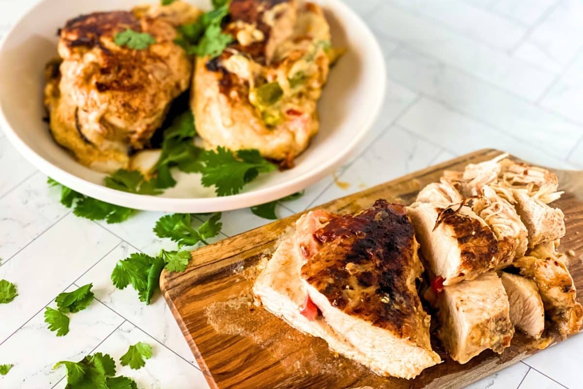 Angled view of a serving dish holding two Fajita Stuffed Chicken Breasts behind a cutting board topped with a sliced chicken breast with juice spilling out of a cheesy center. Hostess At Heart