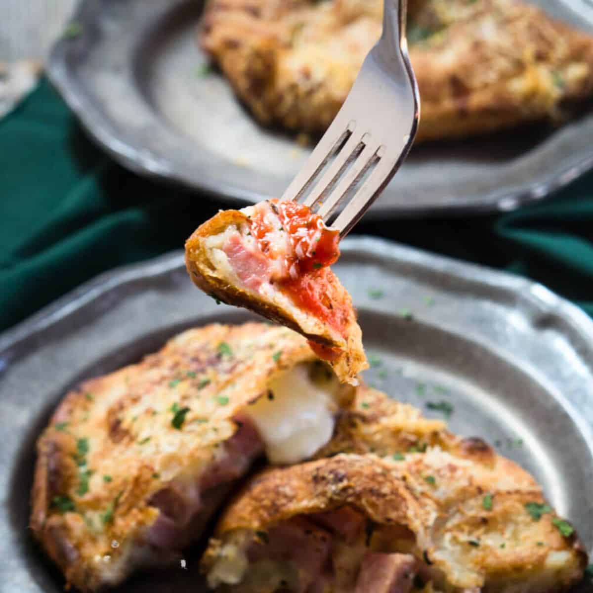 A fork filled bite of a golden baked calzone bite showing juicy ham, melted cheese dipped in marinara sauce. Hostess At Heart