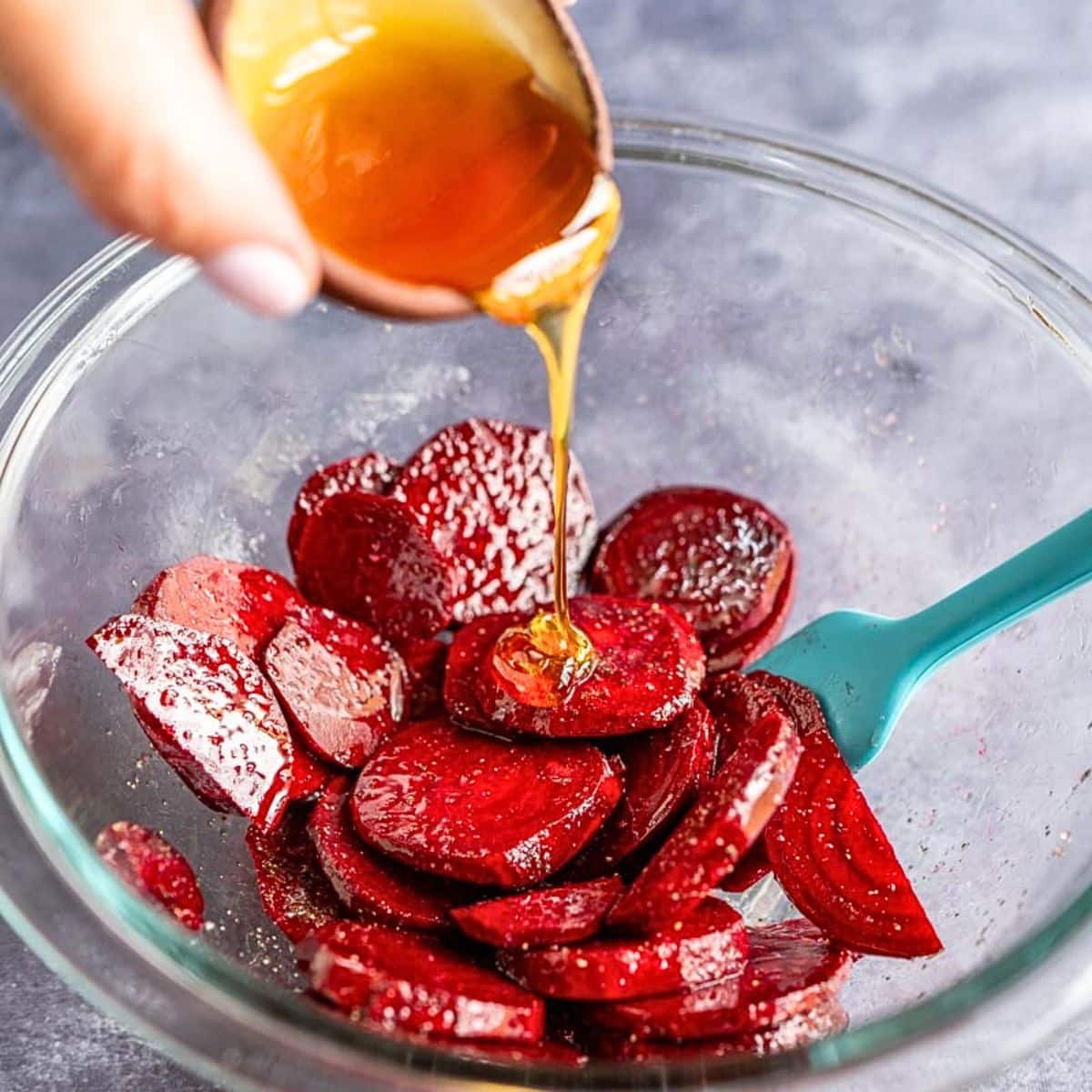 A hand drizzling honey over sliced beets in a mixing bowl inorder to make honey roasted beets. Hostess At Heart