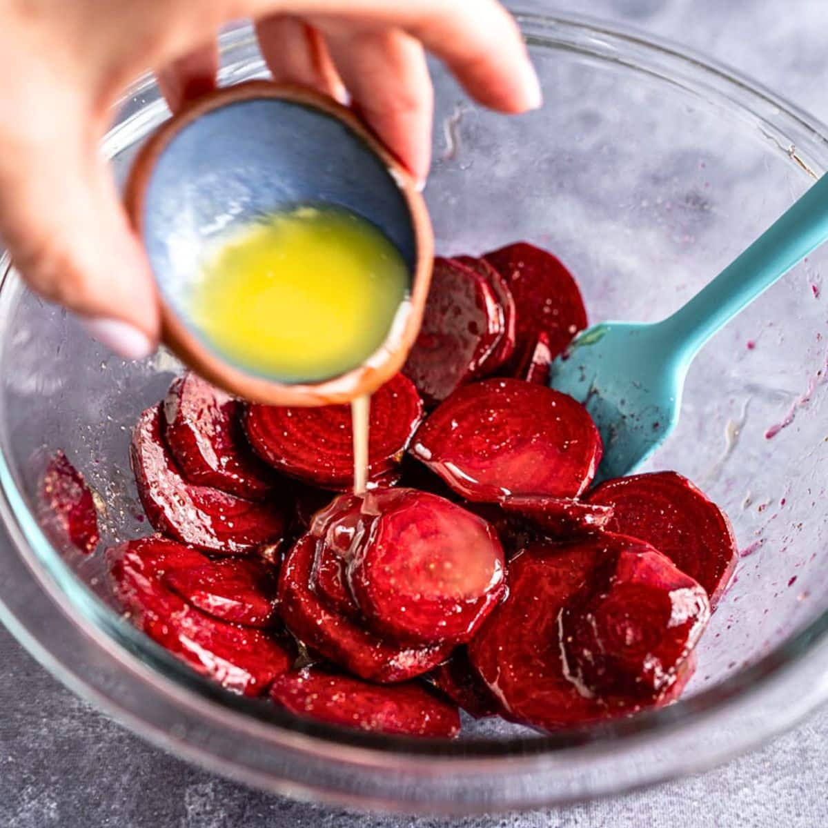 A hand drizzling melted butter over sliced beets in a mixing bowl to make a roasted beets recipe. Hostess At Heart