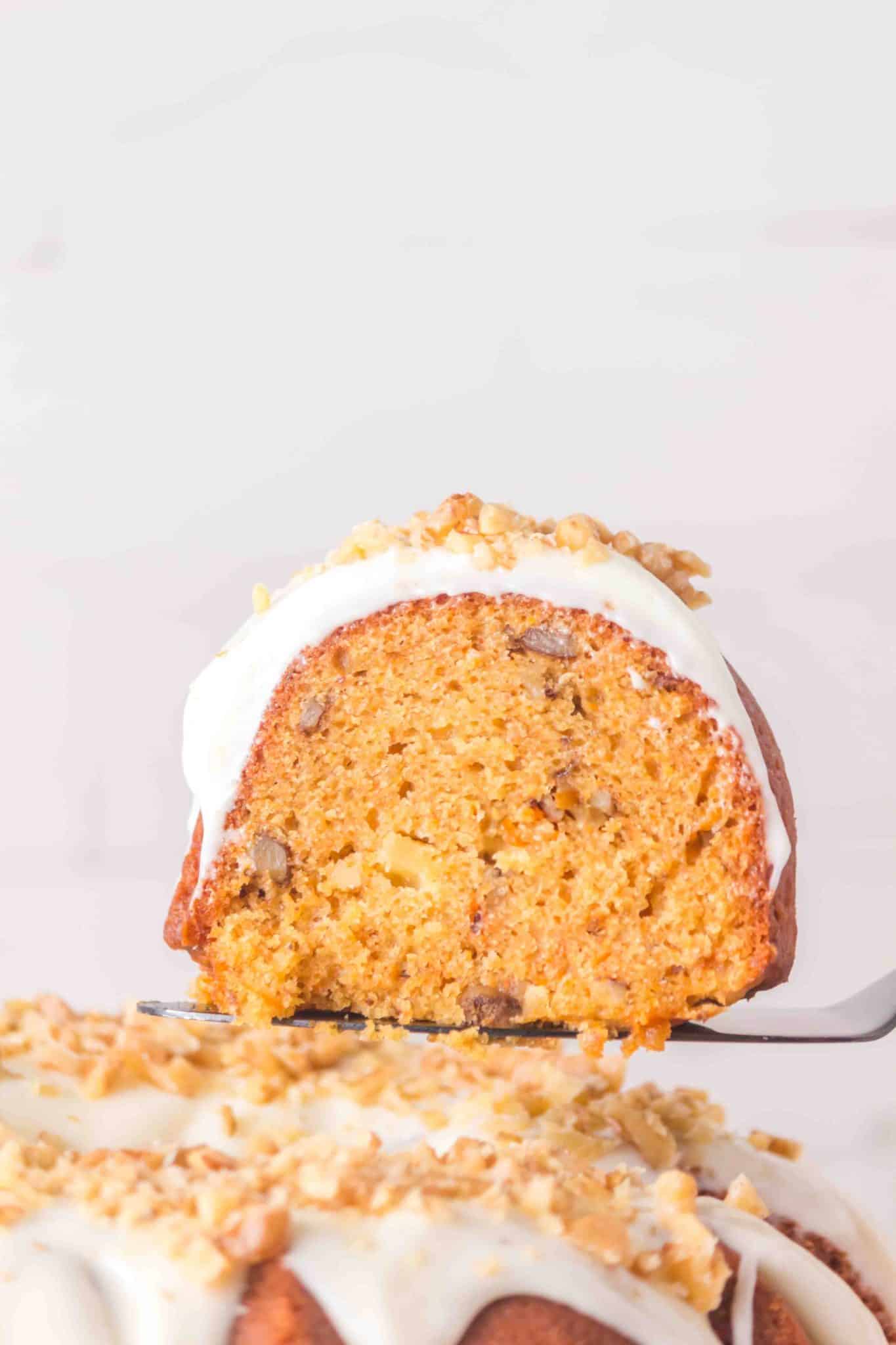 Sideview of a spatula holding a slice of homemade carrot cake showing a moist interior. It's hovering over the bundt cake. Hostess At Heart