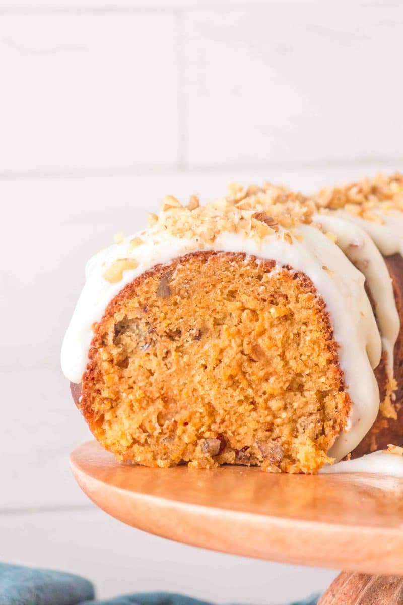 Sliced moist carrot bundt cake on a cake stand showing a tender crumb. It's topped with cream cheese glaze garnished with crushed walnuts - Hostess At Heart