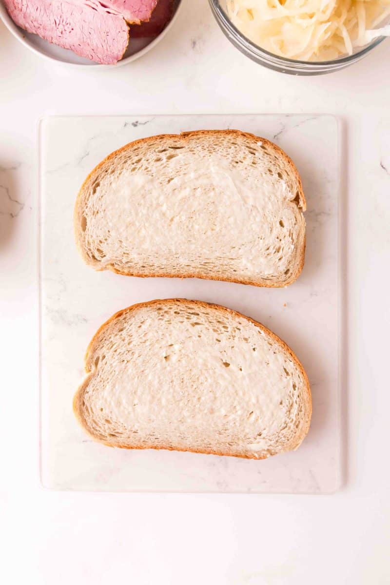 Top down view of two slices of light rye bread slathered with butter for grilled corned beef reuben sandwiches. Hostess At Heart