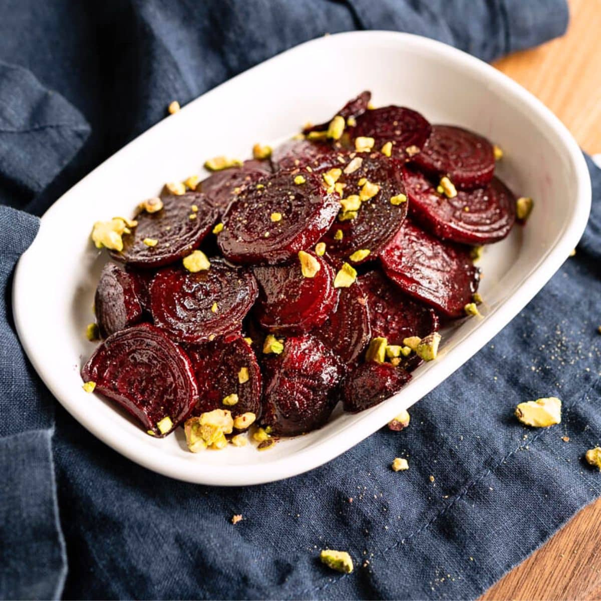 Angled view of roasted beets in a white serving dish glistening from honey and garnished with chopped pistachios. Hostess At Heart