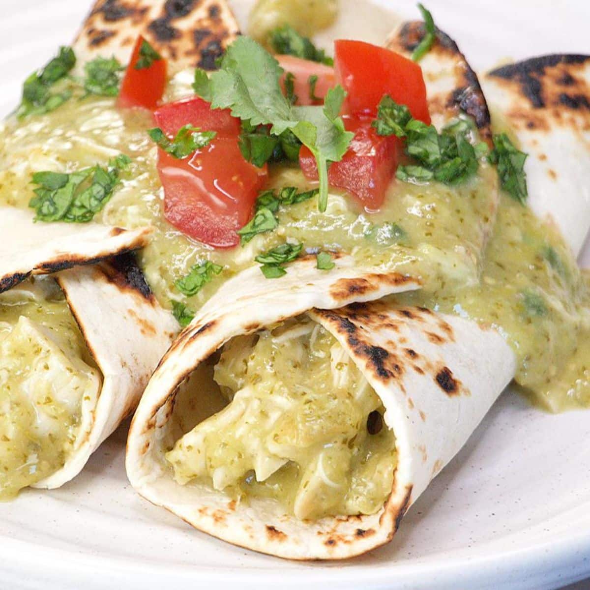 Two green chicken enchiladas on a gray plate smothered in homemade tomatillo mole sauce and garnished with fresh cilantro and chopped tomatoes. Hostess At Heart