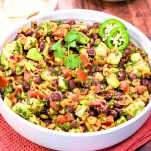 Fresh avocado salsa with corn and black beans in a white bowl next to a bowl of tortilla chips. Hostess At Heart