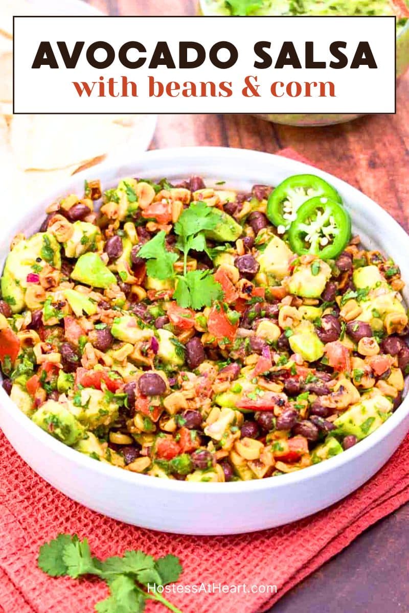 A titled image for Pinterest of Fresh avocado salsa with corn and black beans in a white bowl garnished with cilantro and sliced jalapenos. Hostess At Heart