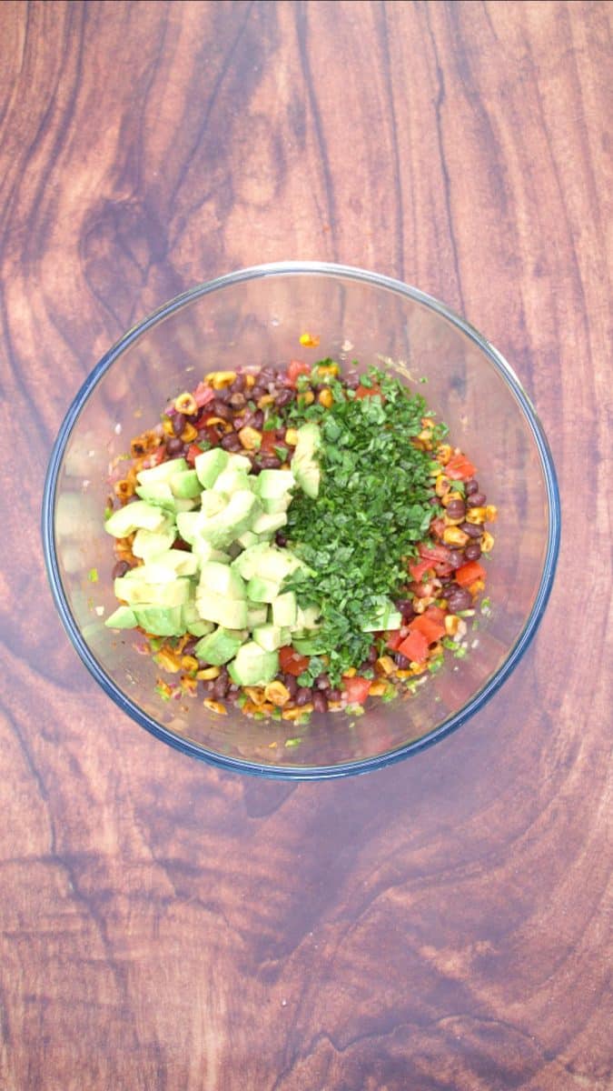 Fresh diced avocado and cilantro added to a mixing bowl filled with a black bean and corn salsa. Hostess At Heart