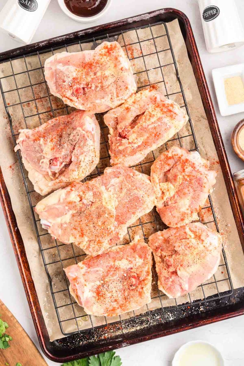 Raw bone-in chicken thighs being coated with dry rub on a parchment-lined baking sheet with rack. Hostess at Heart
