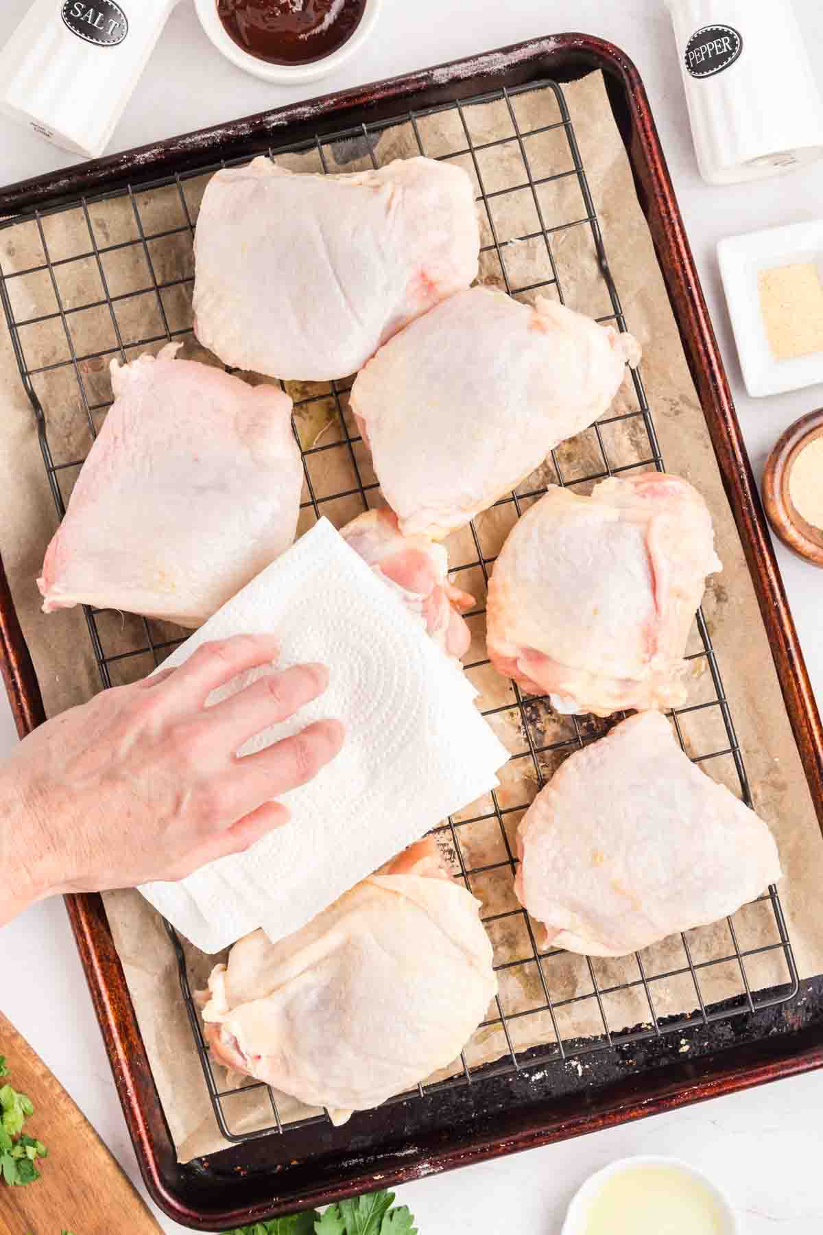 Raw bone-in skin-on chicken thighs on a baking sheet rack being tapped dry with a paper towel before seasoning. Hostess At Heart