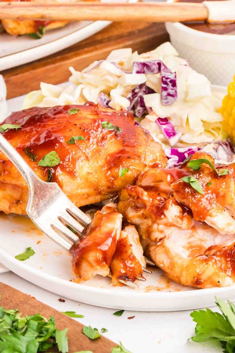 Easy baked barbecue chicken thighs served on a white plate with coleslaw and corn on the cob for a summer dinner. A fork loaded with the juicy chicken is ready to be eaten.