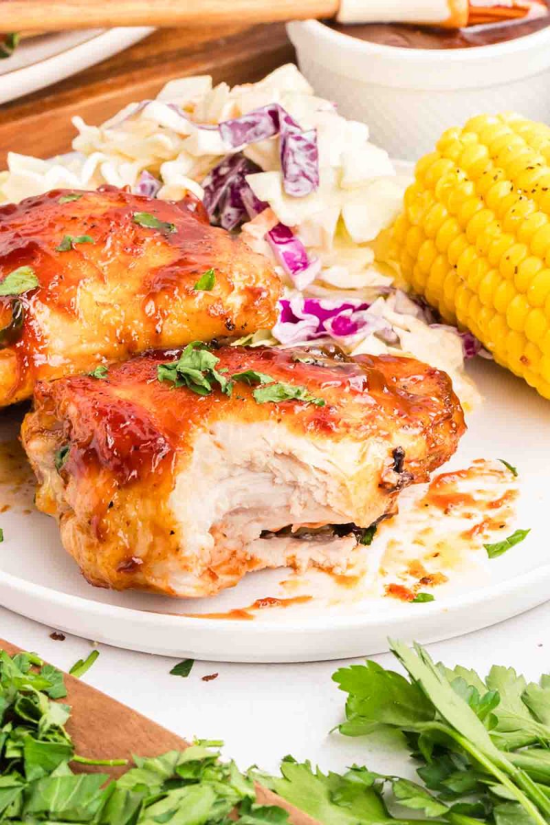 A bite taken out of a baked barbecue chicken thigh showing tender juicy meet and a thick caramelized barbecue sauce on the skin. Hostess At Heart