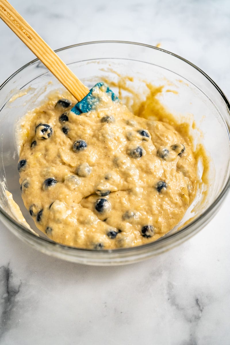 A mixing bowl filled with blueberry banana muffin batter dotted with fresh blueberries. Hostess At Heart