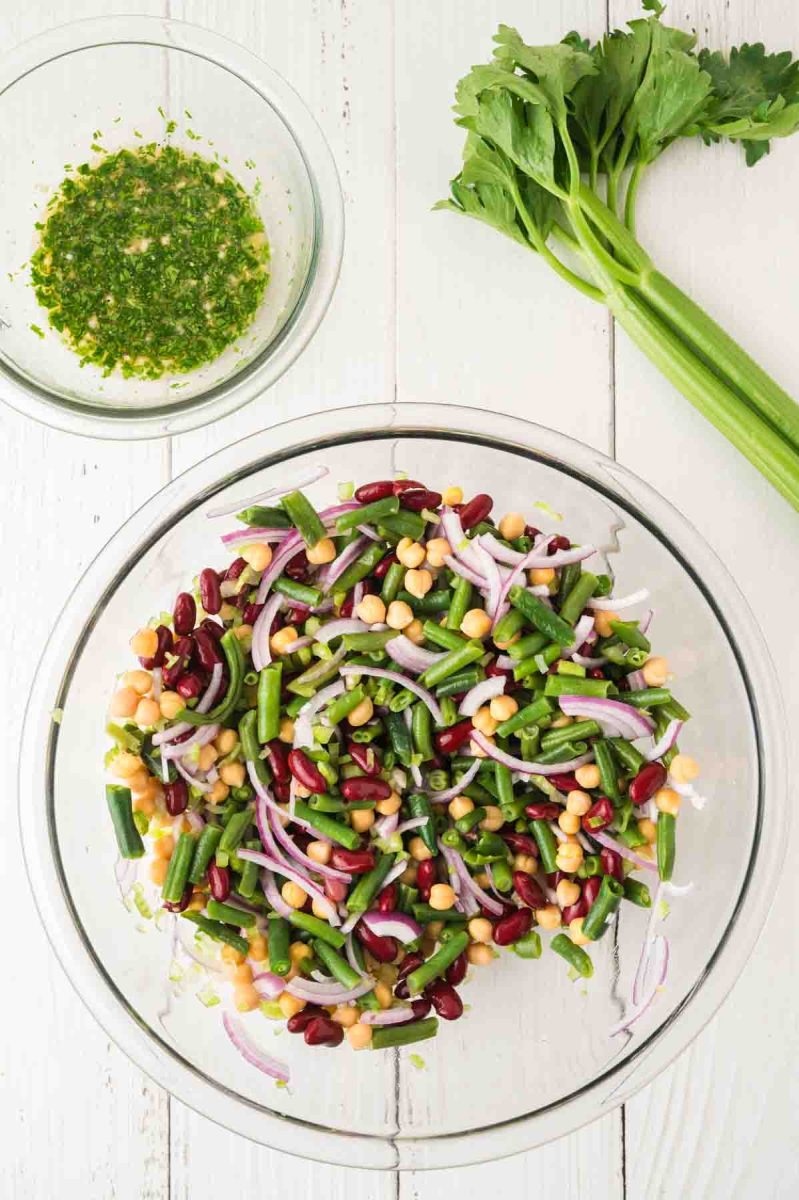 A mixing bowl filled with an undressed cold three bean summer salad with chickpeas, kidney beans, green beans, and red onion. Hostess At Heart