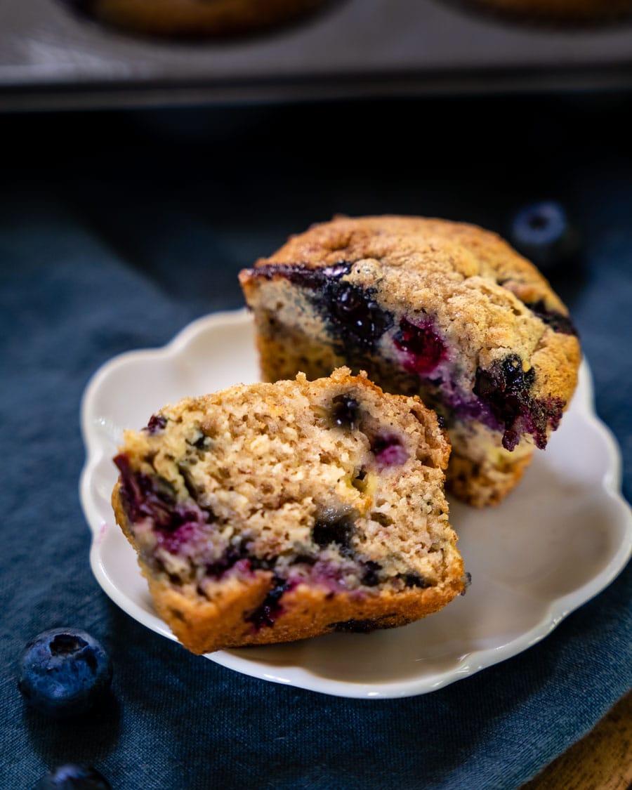A soft and tender banana blueberry muffin cut in half showing purple blueberries throughout the muffin. Hostess At Heart