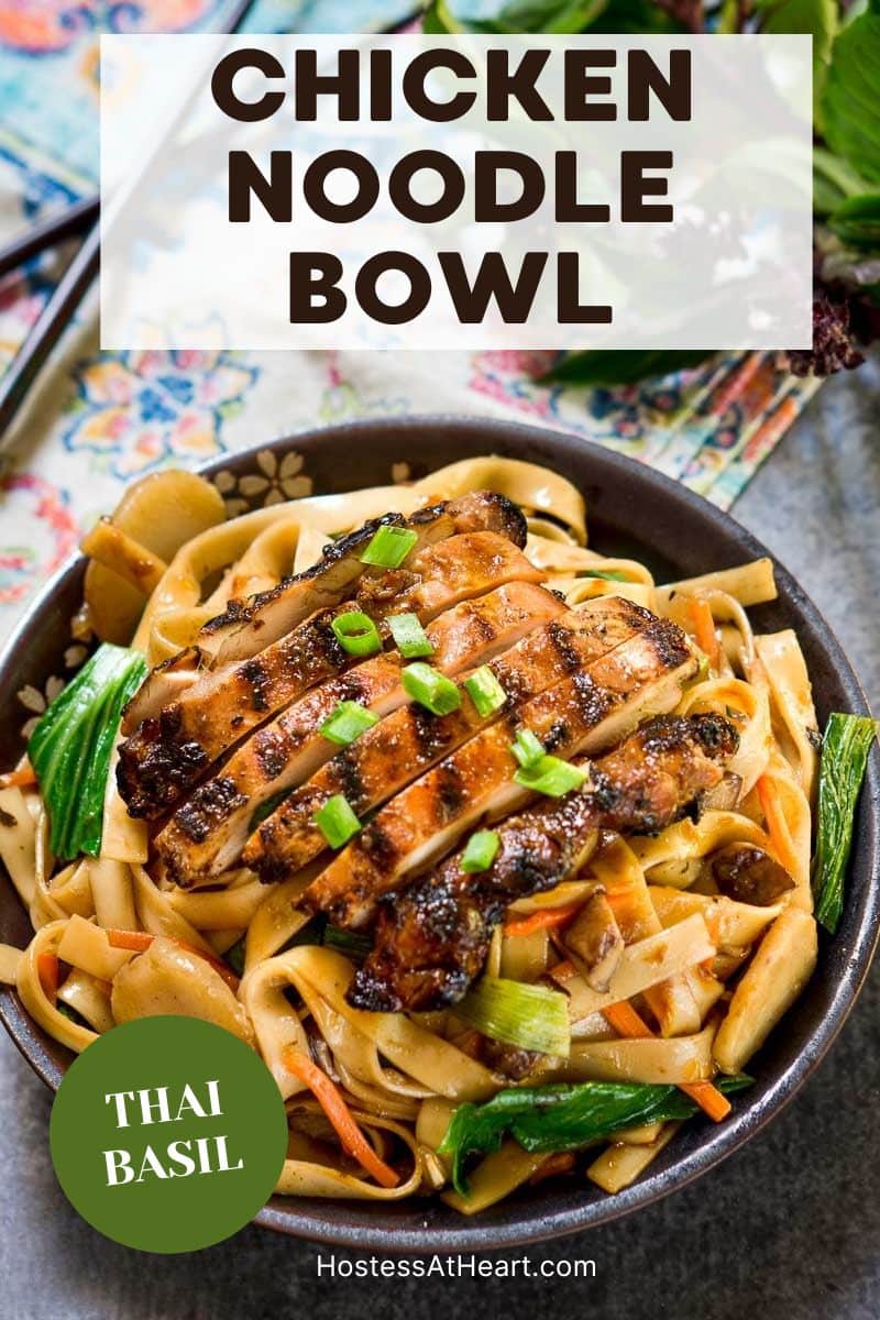 An image for Pinterest of Thai basil noodles in a dark bowl topped with sliced grilled chicken and garnished with green onions and fresh Thai basil. Hostess At Heart
