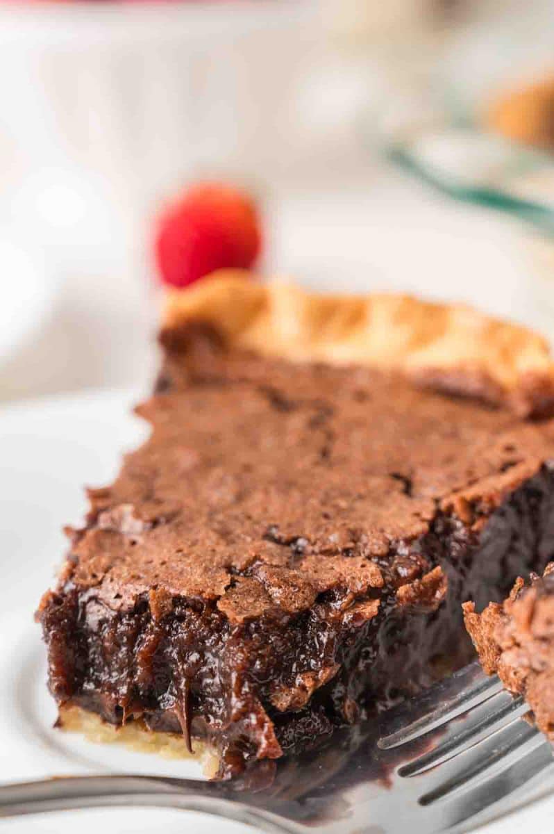 A slice of fudgy chocolate pie from scratch. A fork has removed the first bite showing a gooey fudgy center served on a white plate. Hostess At Heart
