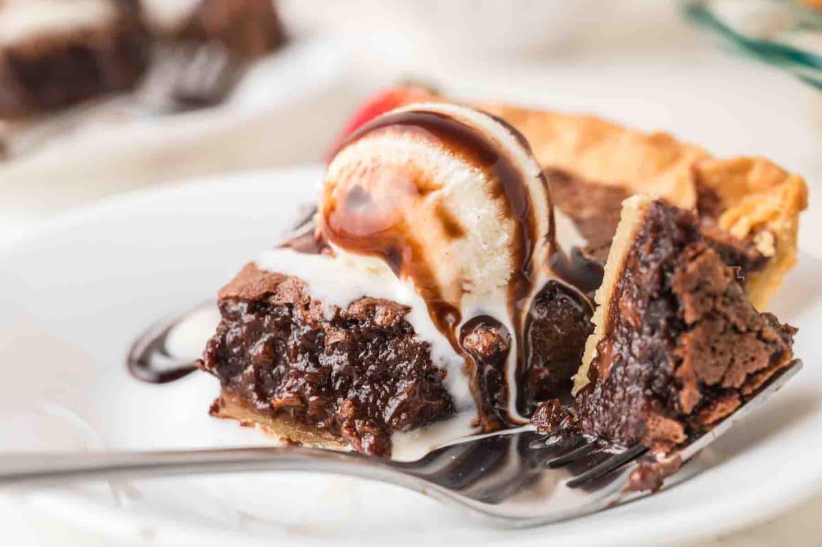 A slice of hot fudge pie with the first bite lifted with a fork showing a gooey fudgy center served on a white plate with a spoon of vanilla ice cream drizzled with chocolate sauce. Hostess At Heart