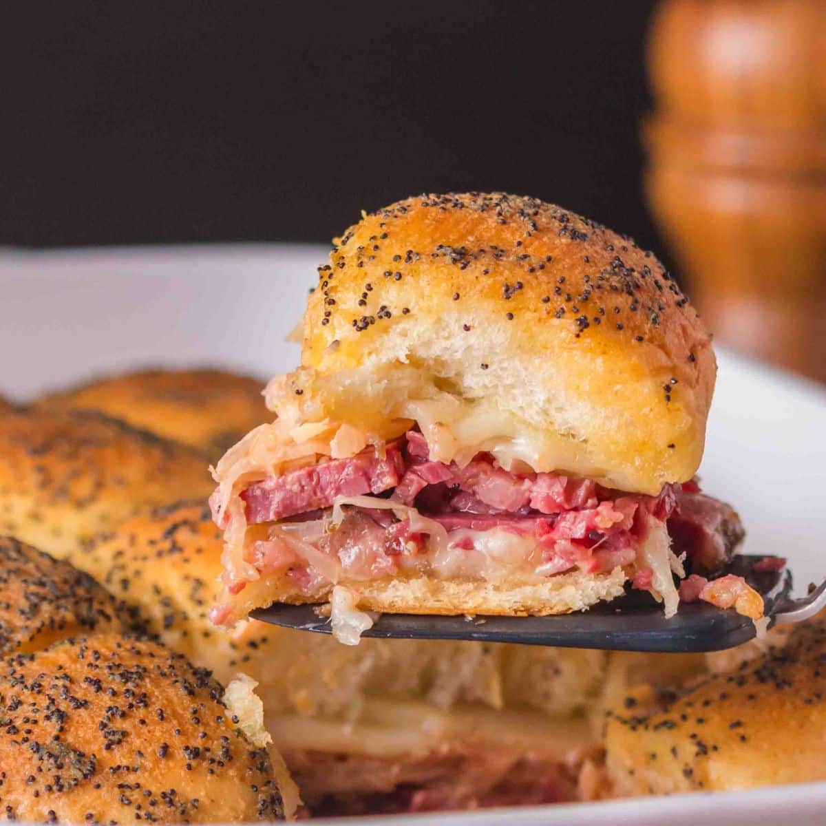 Sideview of a baked slider lifted with a spatula showing layers of juicy corned beef, melted Swiss cheese, and sauerkraut on a golden brown homemade Hawaiian roll. Hostess At Heart