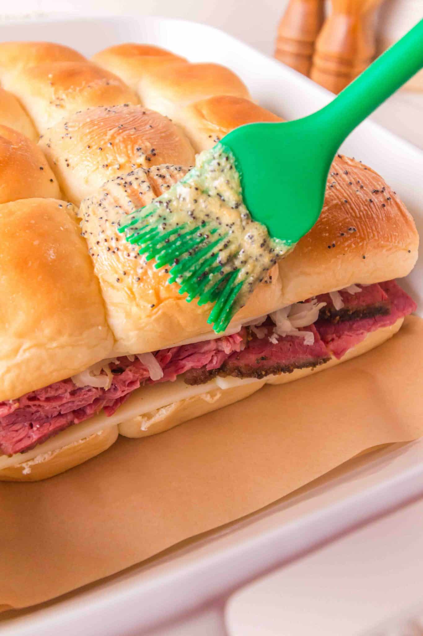 A pastry brush spreading softened poppyseed butter over the top of Hawaiian rolls before baking for corned beef sliders. Hostess At Heart