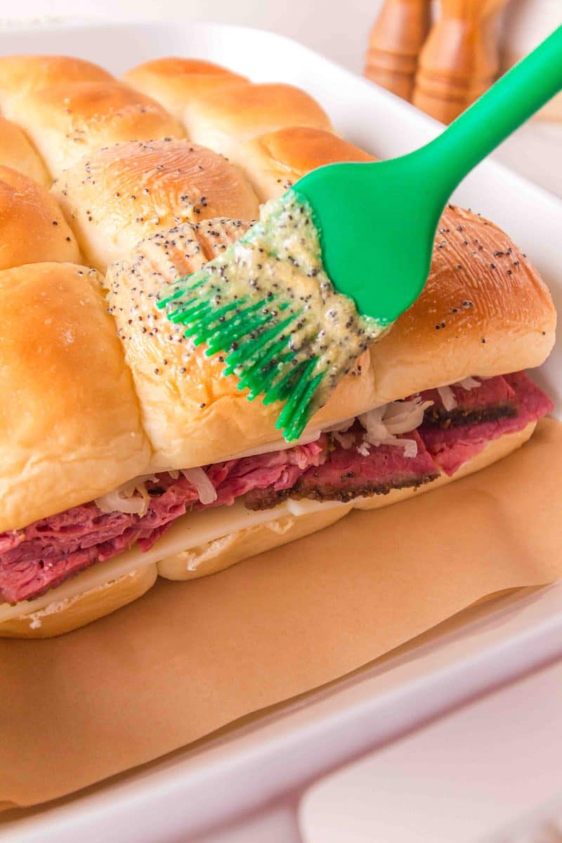 A pastry brush spreading softened poppyseed butter over the top of Hawaiian rolls before baking for corned beef sliders. Hostess At Heart