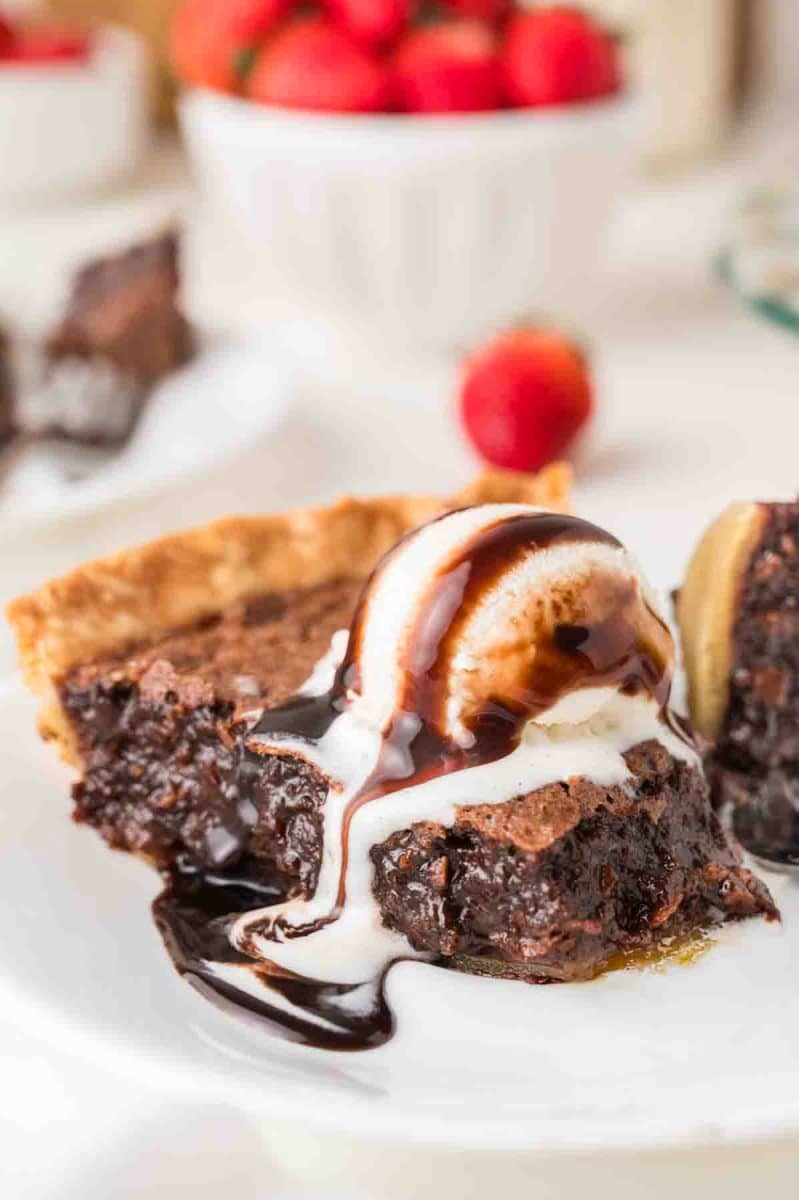 Side view of a slice of chocolate fudge pie with a gooey fudgy center served on a white plate with a scoop of vanilla ice cream drizzled with chocolate sauce. Hostess At Heart