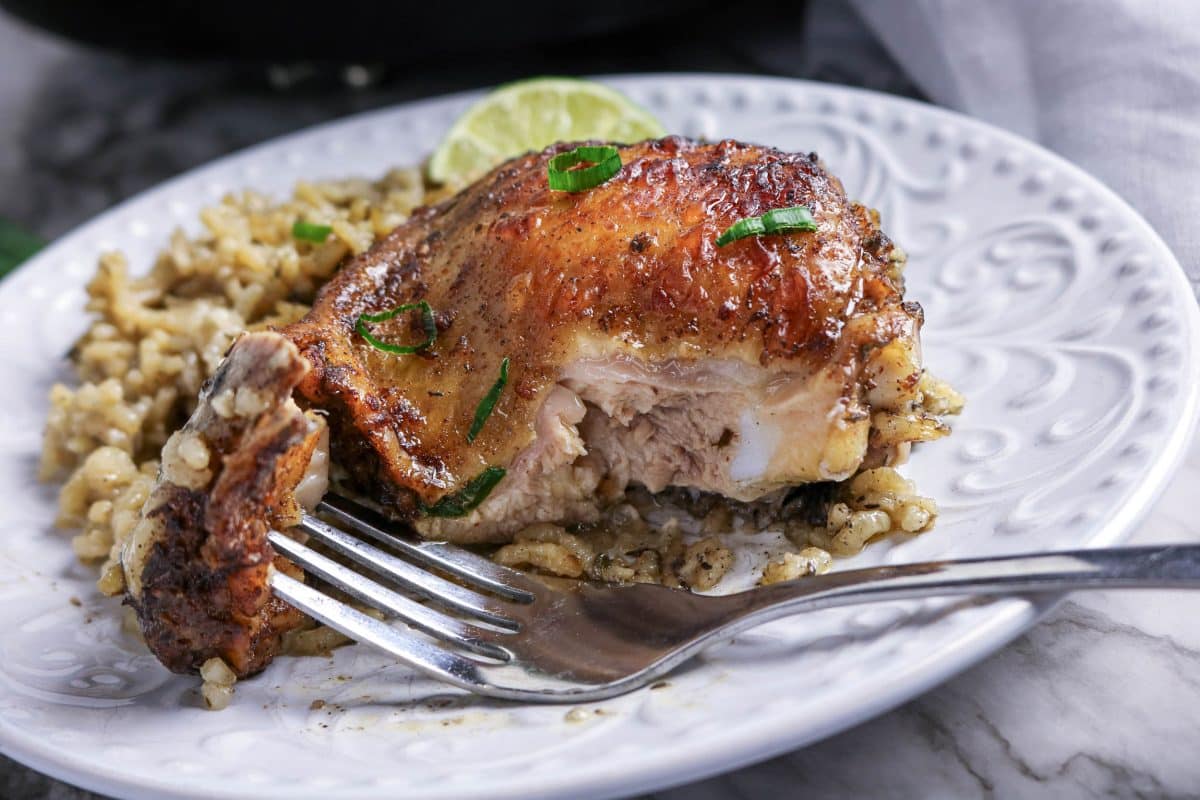 Sideview of a juicy baked jerk chicken though with a fork holding the next spicy bite of jerk chicken on a white plate next to a side of creamy coconut rice. Hostess At Heart