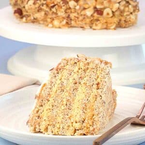 Side view of a slice of Esterhazy cake showing layers of hazelnut meringue and buttercream and the whole Esterhazy Torte on a cake plate behind the slice. Hostess At Heart