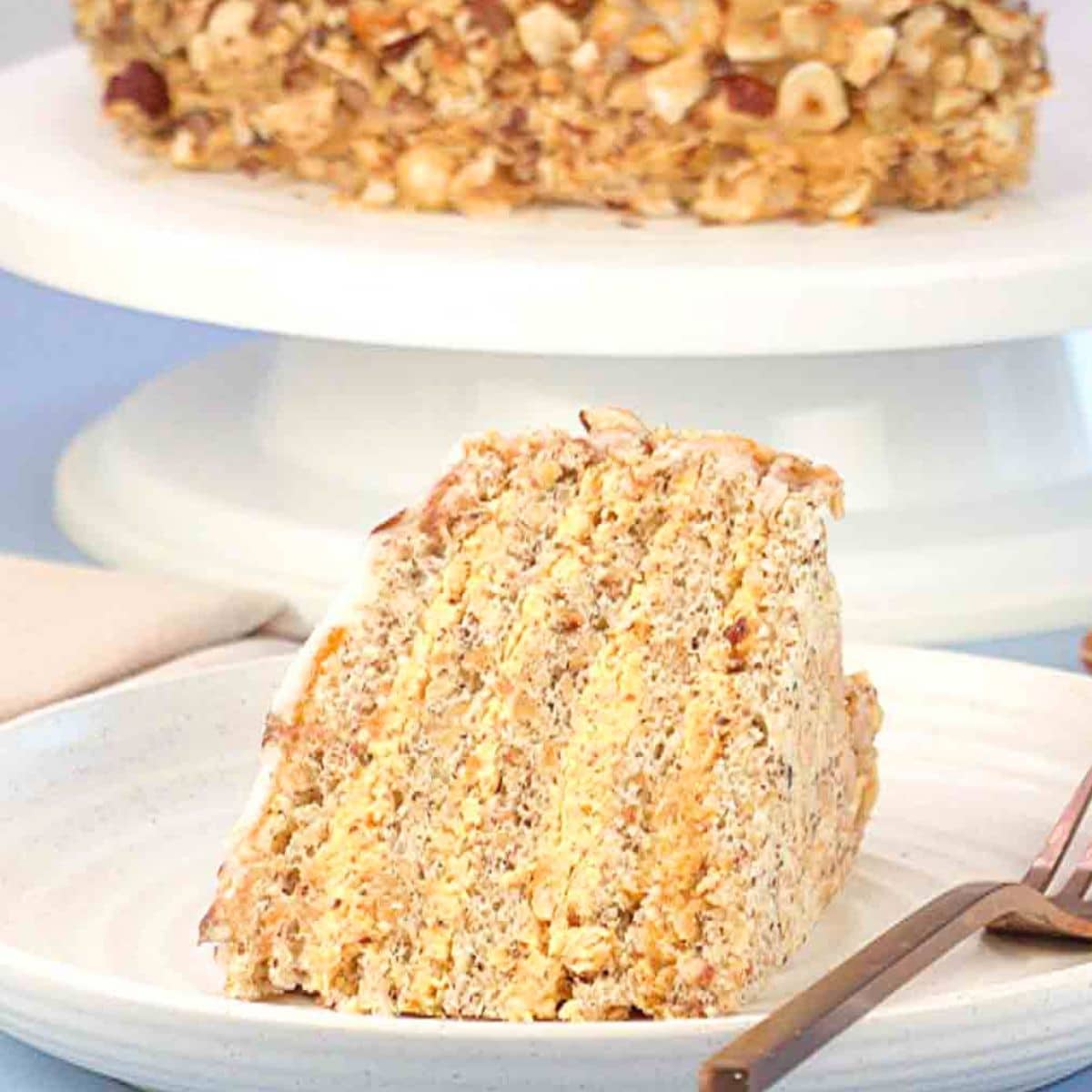 Side view of a slice of Esterhazy cake showing layers of hazelnut meringue and buttercream and the whole Esterhazy Torte on a cake plate behind the slice. Hostess At Heart