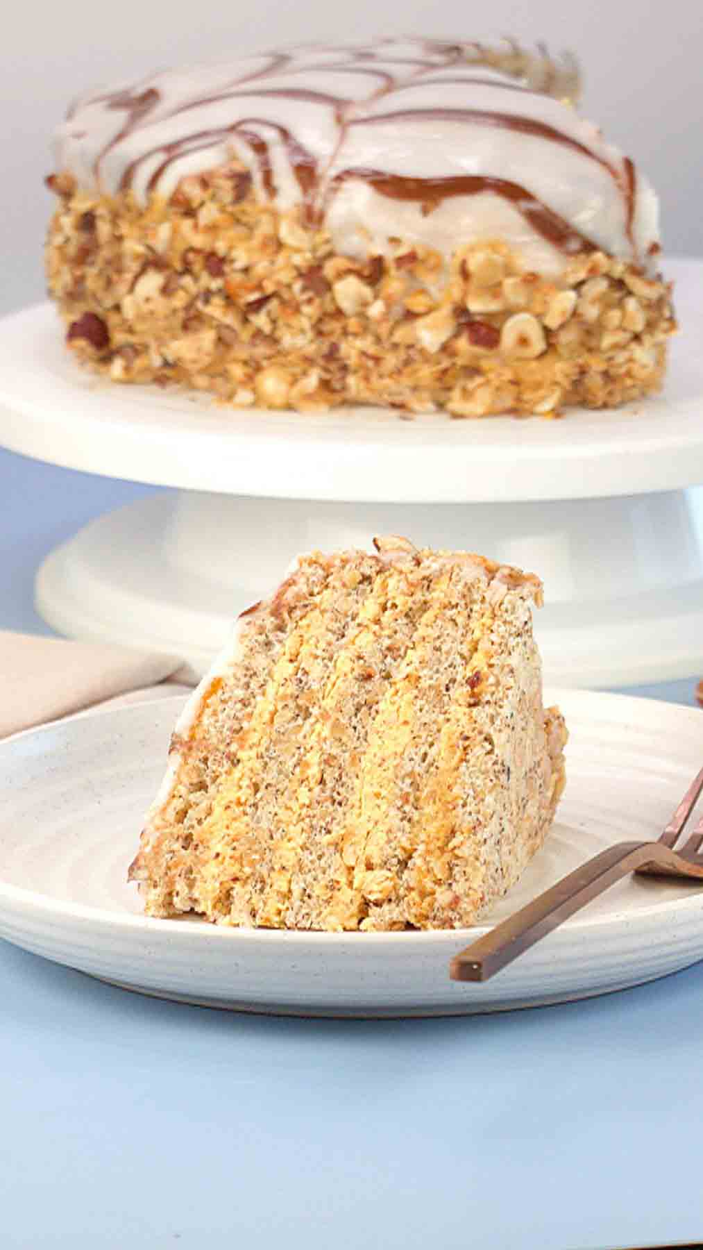 Side view of a slice of Esterhazy cake showing layers of hazelnut meringue and buttercream and the whole Esterhazy Torte on a cake plate behind the slice. Hostess At Heart