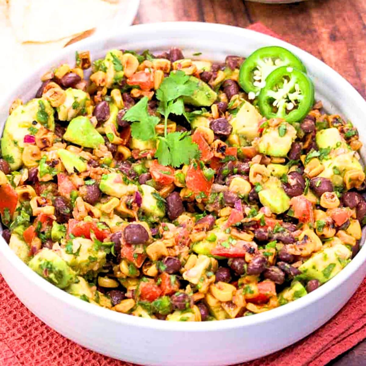 Fresh avocado salsa with corn and black beans in a white bowl next to a bowl of tortilla chips for a no-cook appetizer. Hostess At Heart