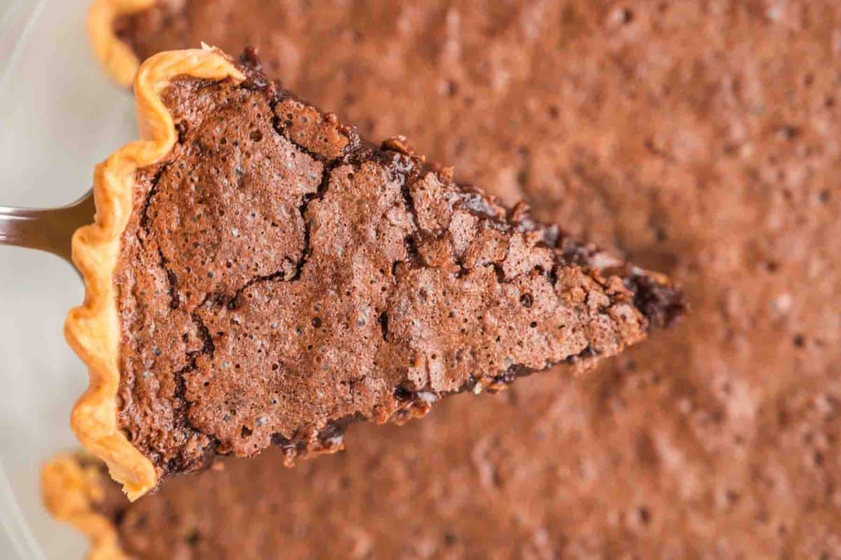 Top down view of a slice of hot fudge pie with a flaky brownie top and golden crust hovering over the whole pie on a pie server. Hostess At Heart