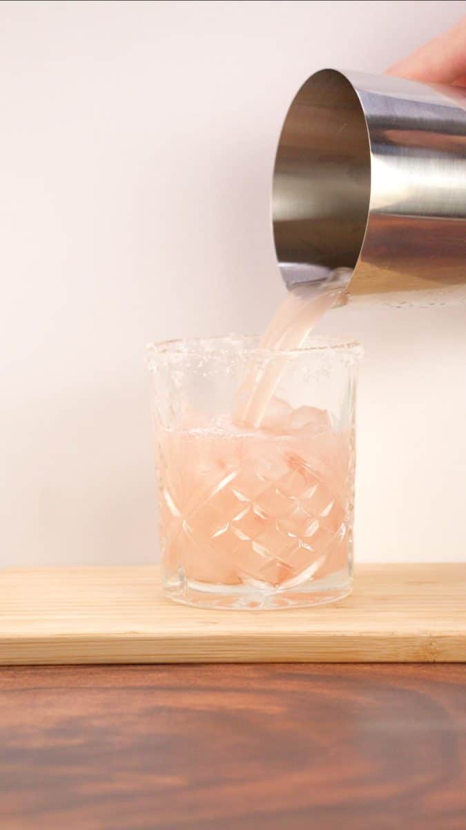 Sideview of a shaker glass pouring a mixed Paloma cocktail into a low rimmed rocks glass. Hostess At Heart