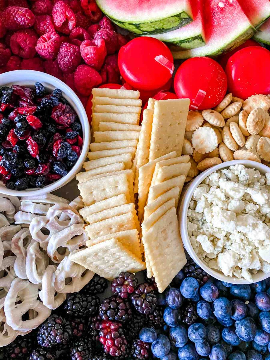Top down view of a board filled with red white and blue charcuterie board ideas including club crackers, blue cheese, babybel cheese, raspberries, blackberries, blueberries, watermelon wedges, and craisins. Hostess At Heart