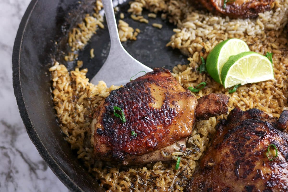 A spatula lifting up a baked jerk chicken thigh from a skillet filled with coconut rice. Hostess At Heart