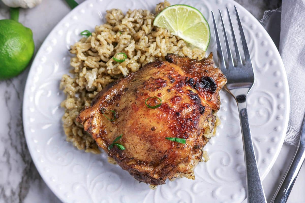 Crispy baked jerk chicken thigh on coconut milk rice with smoky seasoning plated on a white dish garnished with fresh lime wedge and green onions. Hostess At Heart