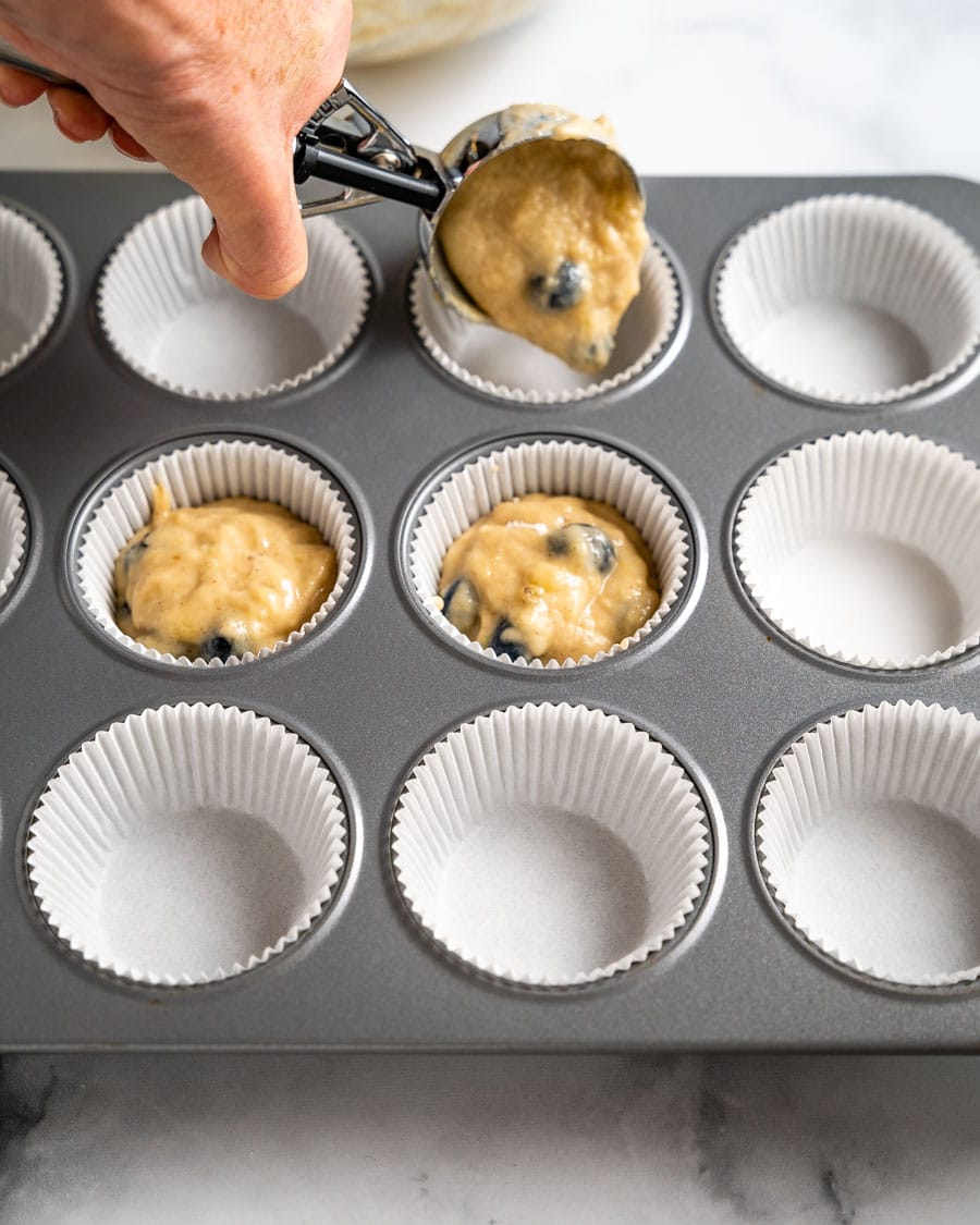 Adding blueberry banana muffin batter to a muffin tin with an ice cream scoop for consistent sizes. Hostess At Heart