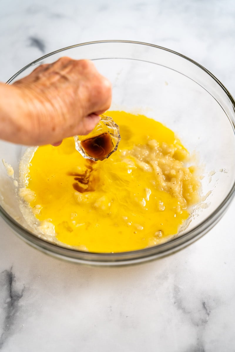 Vanilla extract added to a mixing bowl filled with mashed banana and egg mixture to make blueberry with bananas muffins recipe. Hostess At Heart