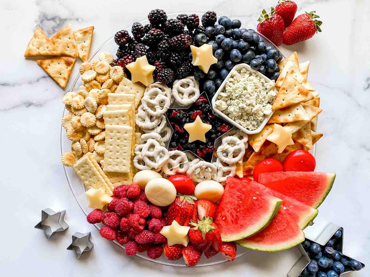 Top down view of a flag charcuterie board featuring fruits, cheeses, crackers, and pretzels in the colors of the American Flag. Hostess At Heart