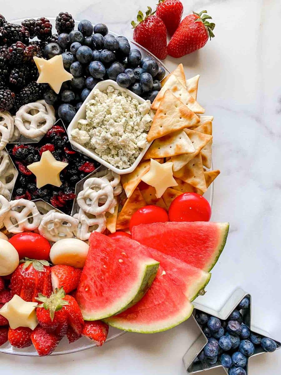 Top down view of a red, white, and blue charcuterie board filled with fruits, crackers, cheeses, and pretzels. Hostess At Heart