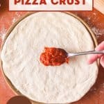Top down view of a spoon filled with pizza sauce getting ready to spread onto a homemade sourdough pie crust made with starter. Hostess At Heart