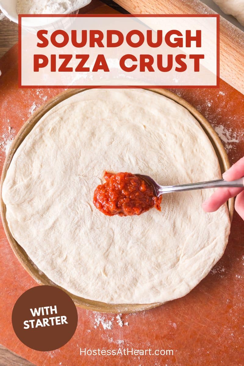 Top down view of a spoon filled with pizza sauce getting ready to spread onto a homemade sourdough pie crust made with starter. Hostess At Heart