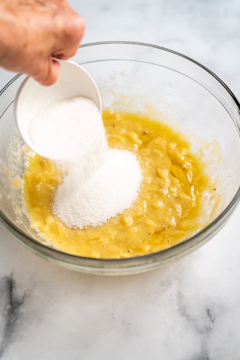 White sugar added to a mixing bowl filled with banana blueberry muffin wet ingredients. Hostess at Heart