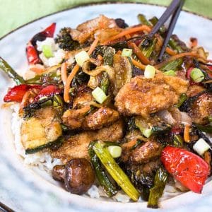 Angled view of a Mongolian chicken stirfry loaded with crispy chicken and beautifully cooked vegetables over a serving of rice on a plate. Hostess At Heart