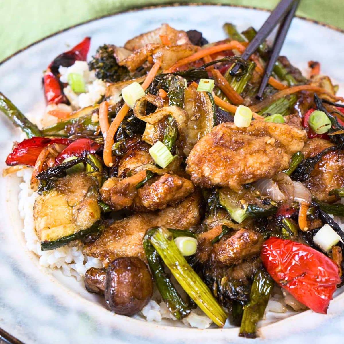 Angled view of a Mongolian chicken stirfry loaded with crispy chicken and beautifully cooked vegetables over a serving of rice on a plate. Hostess At Heart