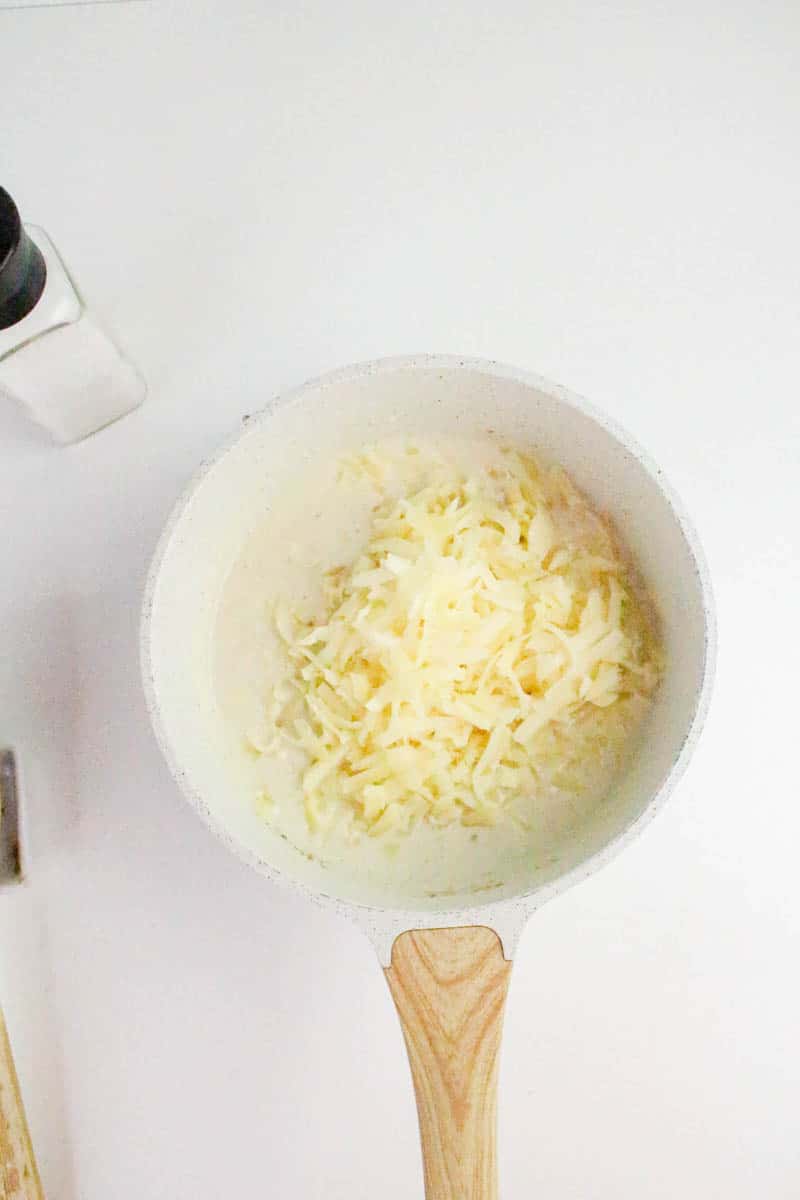 Shredded white cheddar cheese added to simmering cheese sauce in a saucepan. Hostess At Heart